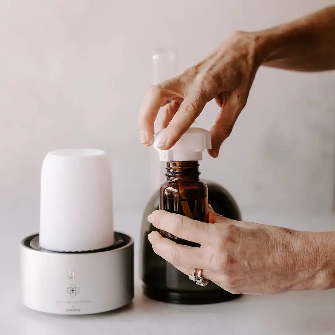 A person’s hands holding a brown glass essential oil bottle with a white cap, next to a silver and white essential oil diffuser.