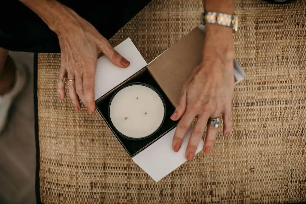 A white, three-wick candle in a black container, nestled in an open box.