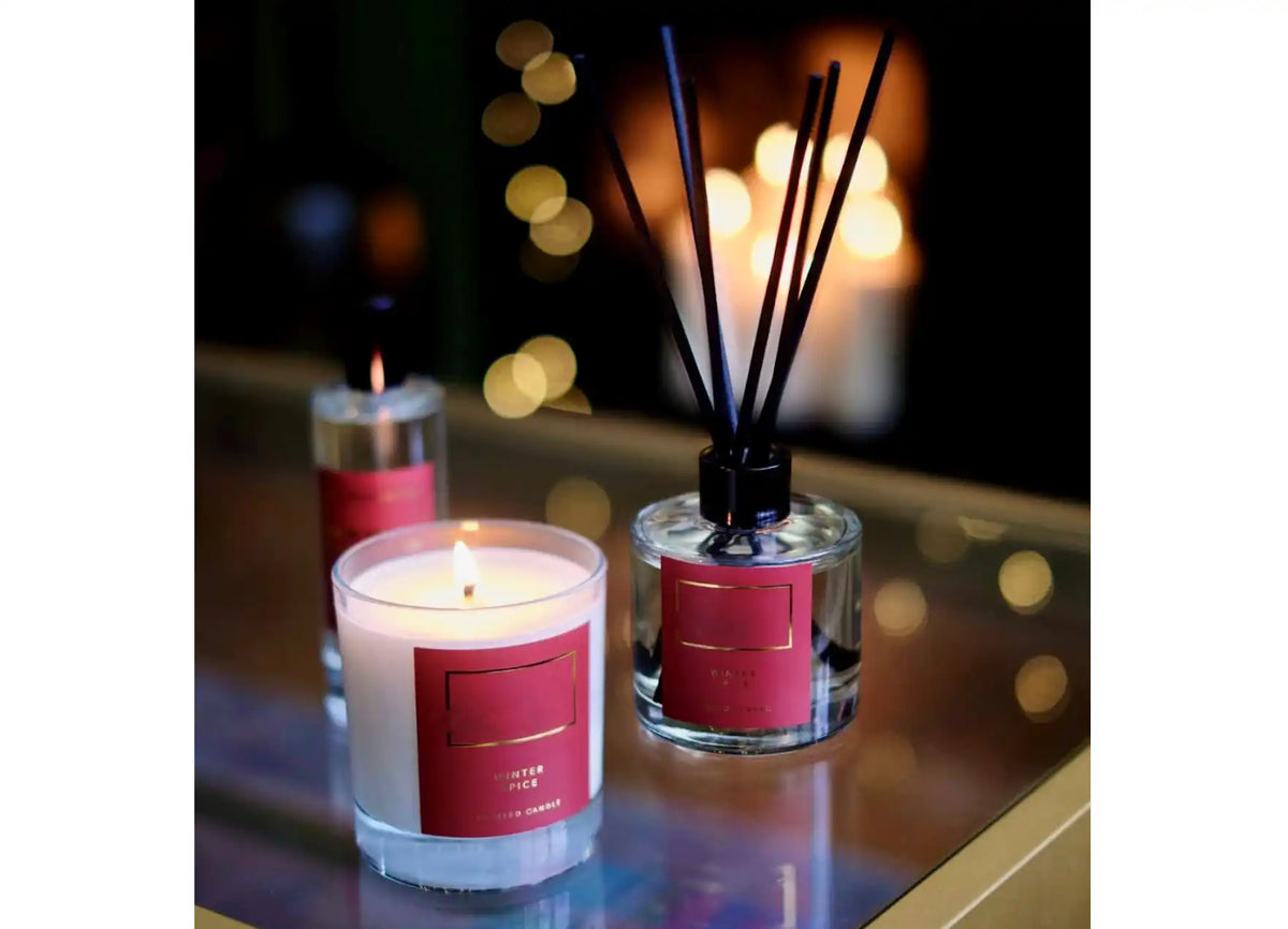 A set of home fragrance products including a lit candle and a reed diffuser, both in clear glass containers with red labels.